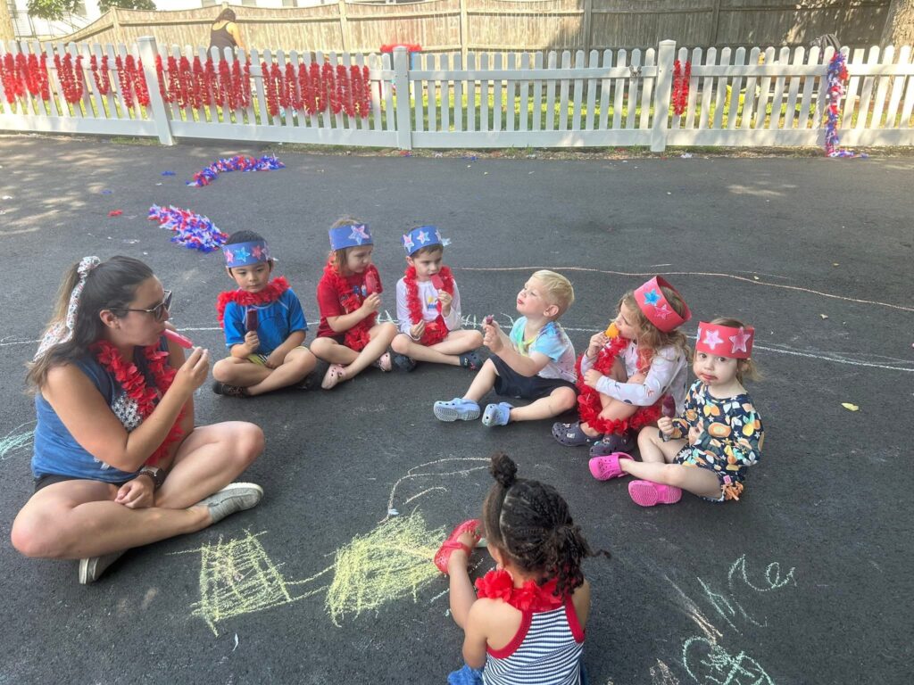Teacher and young children sitting in a circle outside wearing red leis and star crowns while eating popsicles.
